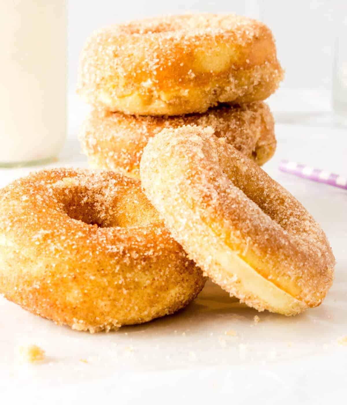 A stack of four homemade donuts coated in cinnamon sugar sits on a white surface, with a bottle of milk and a purple-striped straw blurred in the background.