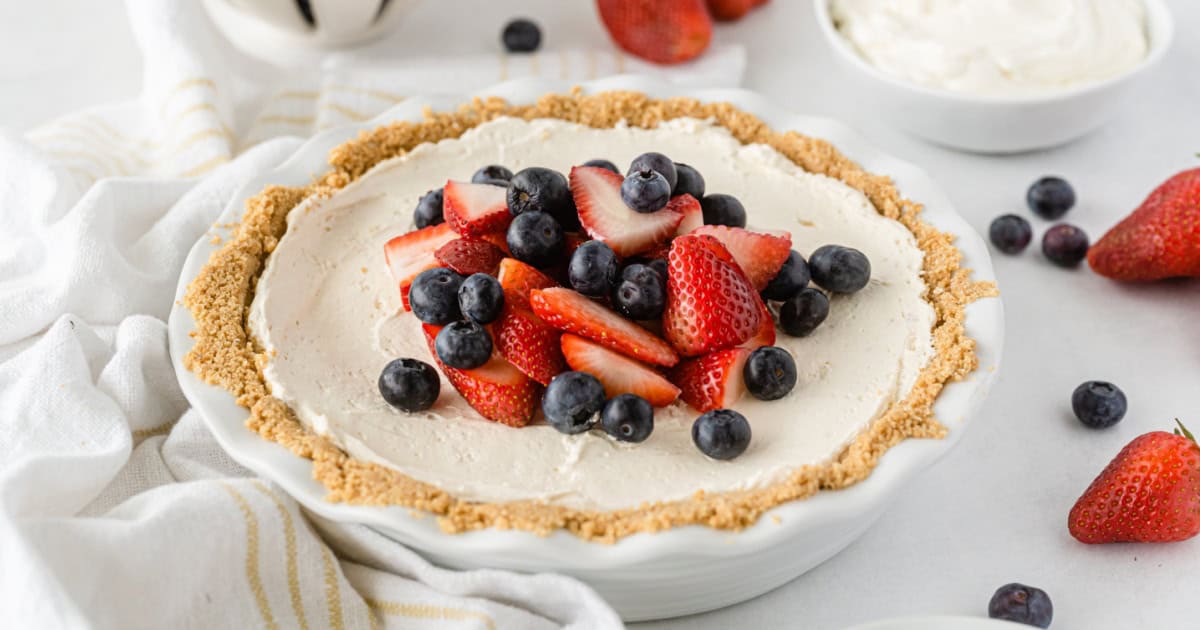 A pie with a crumb crust, creamy filling, and topped with fresh strawberries and blueberries—just like your favorite Cool Whip Desserts. Whole berries and a bowl of cream are nearby on a white surface.
