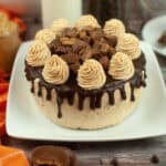 A round Reese’s peanut butter chocolate cake topped with chocolate ganache, peanut butter frosting swirls, and pieces of peanut butter cups, displayed on a white plate with an orange cloth and peanut butter cups nearby.