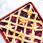 A rectangular berry pie on a red baking sheet, topped with a golden lattice crust and star-shaped pastry cutouts, sits on a white marble surface—perfect for those who love homemade berry pies.