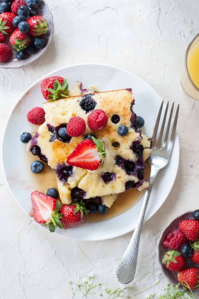 2 squares of pancakes with blueberries and strawberries on a plate, with more fruit scattered around.