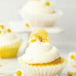 An Air Fryer Cupcake with white frosting, topped with a banana chip and a small daisy flower, sits in a white wrapper. More similarly decorated cupcakes and daisies are in the blurred background.