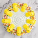 A round Easter Rice Krispie cake topped with white frosting, decorated with yellow marshmallow chicks in a circle, and colorful cereal pieces sprinkled around the edge. The cake sits on a marble surface.