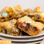 A close-up of colorful Easter blondies with chocolate chunks and pastel candy-coated pieces, stacked on white plates. Some bars have crisp, golden edges and a soft, chewy center. A peach checkered cloth is partially visible in the foreground.