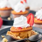 A close-up of a cupcake topped with whipped cream and a sliced strawberry on a black plate. A spoon rests nearby, and other similar cupcakes are blurred in the background. Crumbs are scattered on the plate.