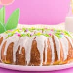 A bundt cake with white icing and colorful sprinkles sits on a white plate. In the background are bottles of milk with straws and decorative felt flowers, making it a charming choice for Easter dessert ideas against a pink backdrop.