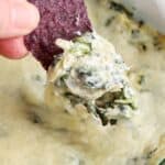 A hand dips a blue corn tortilla chip into creamy spinach artichoke dip, lifting a cheesy portion out of a glass baking dish just like in your favorite spinach artichoke dip recipe.