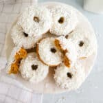 A marble cake stand topped with homemade donuts coated in white icing and shredded coconut; one donut has a bite taken out, revealing a golden brown inside. A checkered cloth and a glass of milk are in the background.