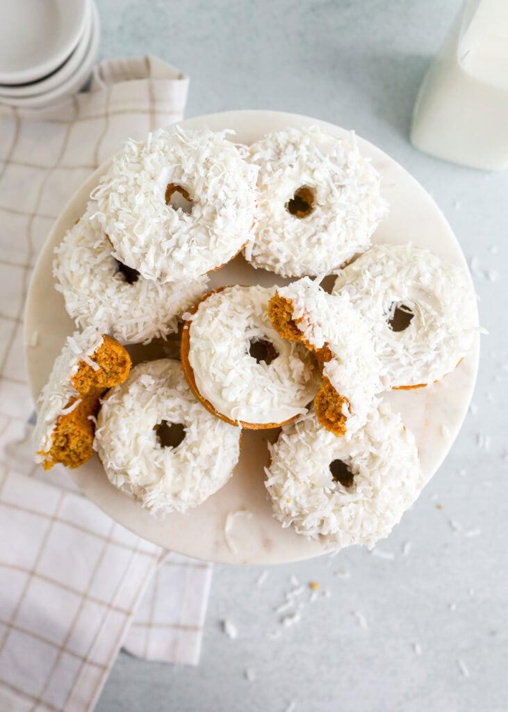 A marble cake stand topped with homemade donuts coated in white icing and shredded coconut; one donut has a bite taken out, revealing a golden brown inside. A checkered cloth and a glass of milk are in the background.