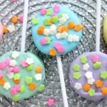Colorful cookie pops covered in pastel icing (pink, blue, green, and purple) are topped with flower-shaped sprinkles and displayed on a woven silver surface—perfect for inspiring your next spring cookie recipes.
