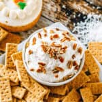 A bowl of carrot cake dip topped with chopped nuts and shredded coconut. Surrounded by graham crackers.