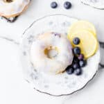 A homemade donut on a floral plate, glazed to perfection and garnished with fresh blueberries and two lemon slices, with more blueberries scattered nearby on a white marble surface.