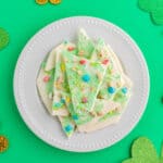 A white plate with pieces of green and white marbled candy bark topped with colorful sprinkles, surrounded by gold chocolate coins and shamrock decorations, makes this a festive choice for St Patrick’s Day Desserts on a bright green background.