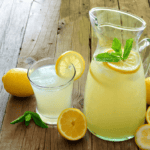 A glass and a pitcher of lemonade, made from a classic lemonade recipe and garnished with lemon slices and mint leaves, sit on a rustic wooden table, surrounded by whole and halved lemons and fresh mint sprigs.