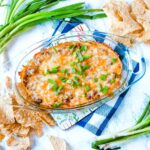 A glass dish of melted chili cheese dip topped with chopped green onions, with tortilla chips and green onions on the side.