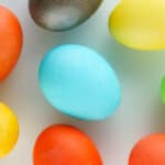 A close-up of nine brightly colored Easter eggs, created using egg dying with Kool-Aid, including orange, yellow, blue, green, and brown eggs arranged randomly on a white background.