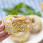 A hand holds a partially eaten glazed donut topped with orange zest. In the blurred background, more donuts—showcasing festive Christmas donut flavors—rest on a plate alongside green leaves and an orange slice.