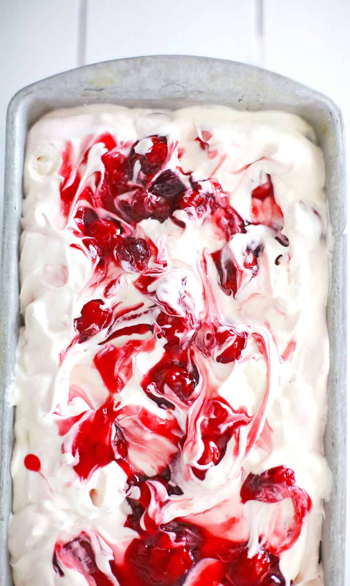 A close-up of cherry cheesecake ice cream in a loaf pan, its creamy white surface marbled with bright red cherry sauce swirls.