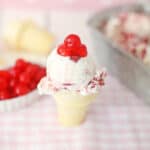 A scoop of cherry Christmas ice cream in a small cone, topped with two maraschino cherries, sits on a pink and white checkered cloth with a tray of cherries and ice cream blurred in the background.