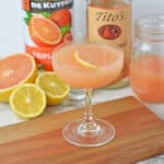 A Grapefruit Martini Recipe shines in a coupe glass with a lemon slice garnish on a wooden board. In the background are halved grapefruit and lemon, pink juice, and bottles of Titos vodka and DeKuyper Triple Sec.
