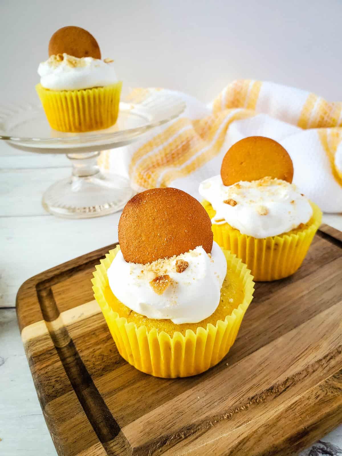 Two Banana Cream Pie Cupcakes in yellow wrappers, topped with whipped cream, a round cookie, and sprinkled crumbs sit on a wooden tray, with a third cupcake in the background on a glass stand. A yellow and white towel is nearby.
