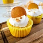 A cupcake in a yellow wrapper, made with yellow cake mix and topped with whipped cream, a vanilla wafer cookie, and cookie crumbs sits on a wooden tray, with another similar cupcake and a striped towel in the background.