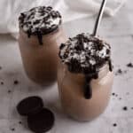 Two mason jars filled with Oreo Milkshake topped with whipped cream, chocolate syrup, and cookie crumbs, with a spoon in one jar. Two chocolate sandwich cookies are placed beside the jars on a marble surface.