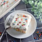 A square piece of frosted cake, inspired by depression era desserts, with colorful sprinkles sits on a white plate with a fork, next to more sprinkles and green leaves, on a tiled surface.