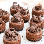 Chocolate Cheesecake Bites topped with chocolate curls are arranged on parchment paper. Each dessert features a chocolate base, creamy filling, and decorative chocolate shavings for an irresistible treat.