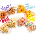 Seven clear butterfly snack bags contain different treats including pretzels, cheese puffs, colorful and plain cereals, each sealed with a colorful twisted pipe cleaner on a white background.