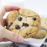 A hand holding a large chocolate chip cookie with melted chocolate chunks—perfect as Mother’s Day cookies—with more cookies and chocolate chips on a plate in the background.