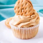 A cupcake with swirled peanut butter frosting is topped with a peanut butter cookie, displayed on a white plate with a light blue cloth—an irresistible pick for those who love dessert recipes with peanut butter.