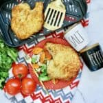 Plate with breaded chicken cutlet on a salad with carrots, next to tomatoes and lettuce. Another cutlet in an air fryer basket with spatulas, plus salt and pepper containers—perfect alongside herbs for pork chops—on a red and gray patterned cloth.