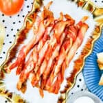 A gold-trimmed dish holds several air fryer bacon twists. Nearby are slices of toasted bread on a blue plate, a gold spatula, a tomato, and a white bowl, all set on a polka-dotted tablecloth.