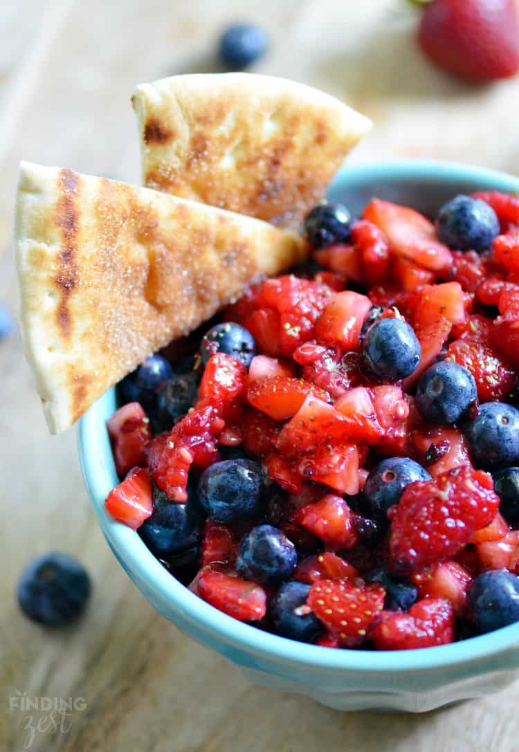 A bowl filled with chopped strawberries and whole blueberries is topped with two cinnamon-sugar pita chips. Perfect for Memorial Day dessert recipes, a whole strawberry and extra berries are scattered on the wooden surface nearby.