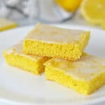 Three lemon bars with a glossy icing are stacked on a white plate. The bars have a bright yellow color and moist, cake-like texture—a perfect example of creative uses for yellow cake mix. A sliced lemon is blurred in the background.