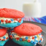 Three patriotic 4th of July Whoopie Pies with red tops, blue bottoms, and white cream filling, decorated with red, white, and blue star sprinkles, are stacked on a black plate with a blurred background.