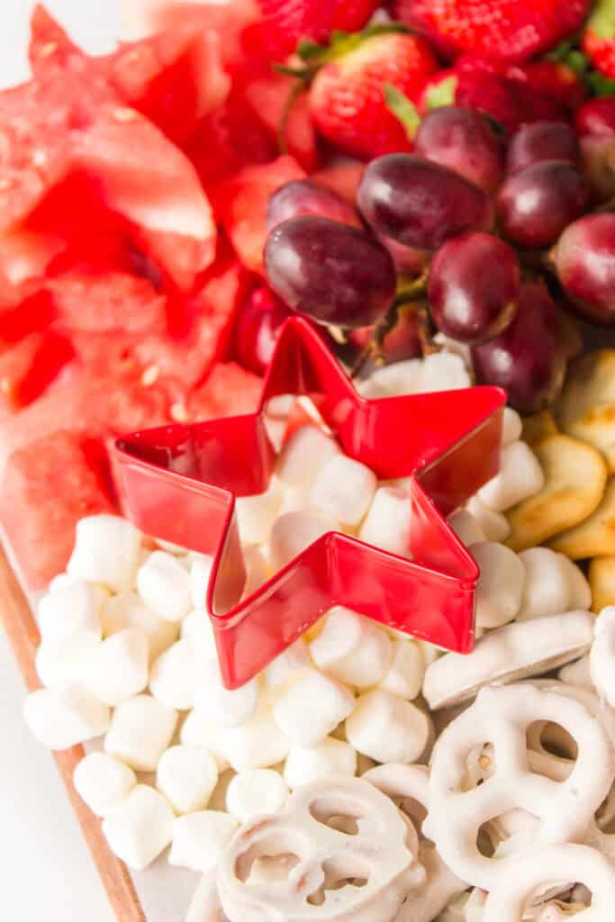 A red star-shaped cookie cutter sits on a platter surrounded by mini marshmallows, yogurt-covered pretzels, watermelon slices, strawberries, and grapes.