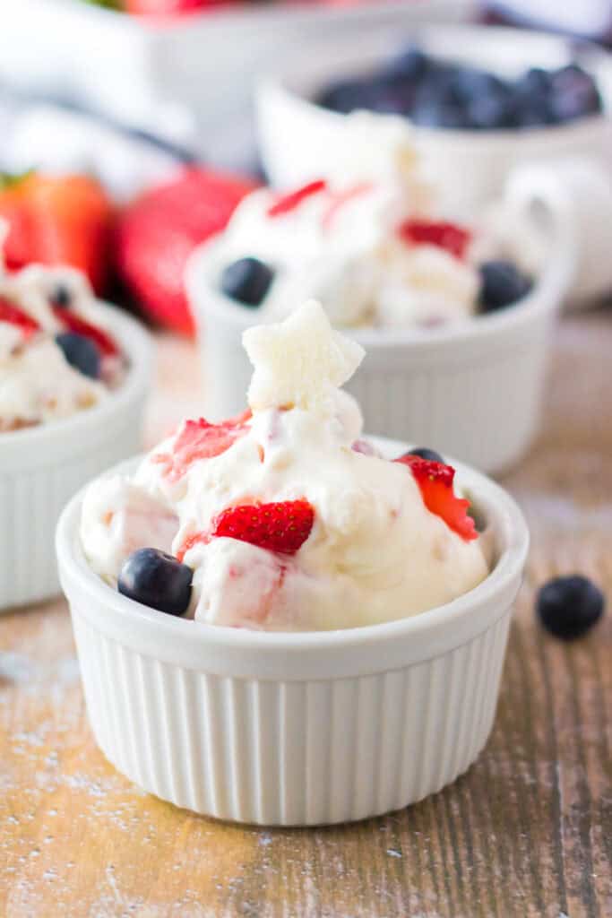A small white ramekin filled with creamy dessert, mixed with fresh strawberries and blueberries, topped with a star-shaped piece of fruit—perfect for Memorial Day Salads. More ramekins and fruits are blurred in the background on a wooden surface.