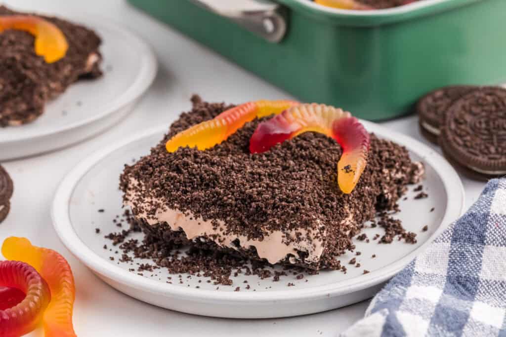 A Cool Whip Desserts favorite, this treat features crushed chocolate cookies, creamy filling, and colorful gummy worms, all served on a white plate with a baking dish and whole cookies in the background.