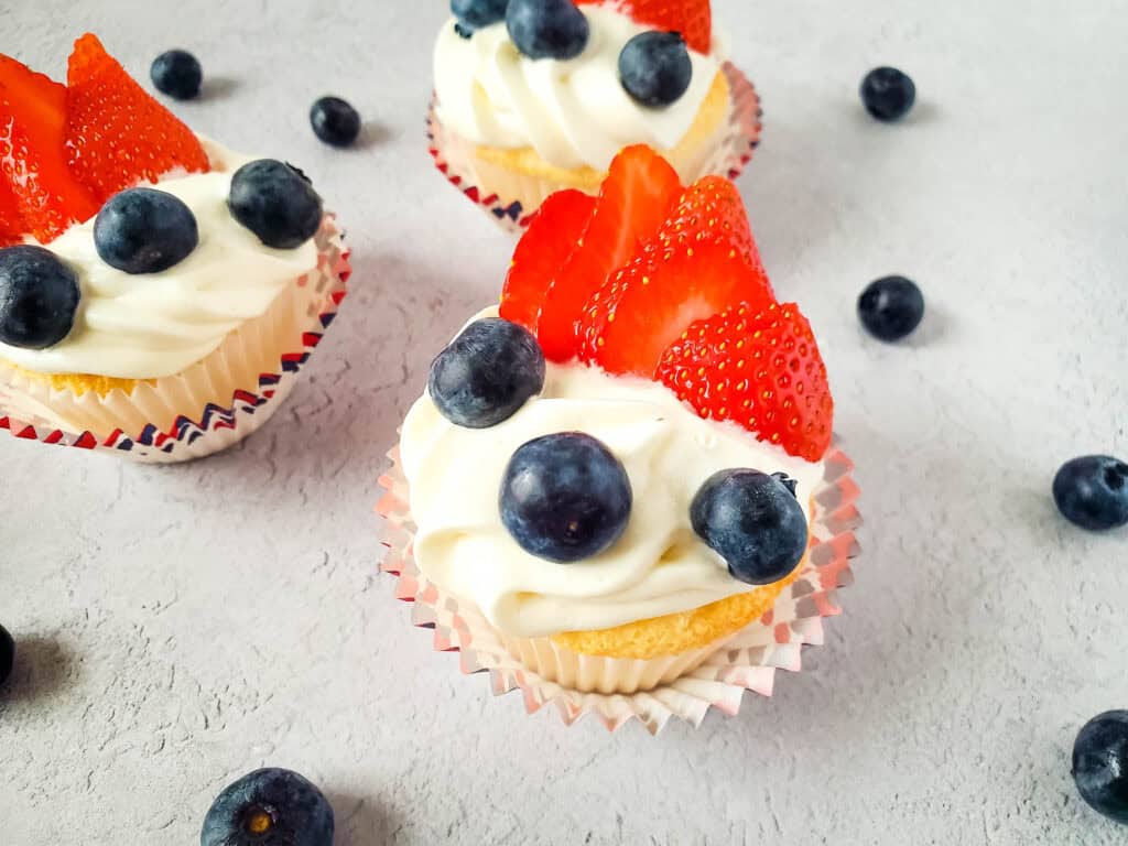 Angel food cupcakes topped with swirls of white frosting, fresh blueberries, and sliced strawberries, arranged on a light gray surface with loose blueberries scattered around.