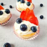 Angel food cupcakes topped with swirls of white frosting, fresh blueberries, and sliced strawberries, arranged on a light gray surface with loose blueberries scattered around.