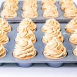 Two trays of Peanut Butter Chocolate Chip Cupcakes with tall swirls of light brown frosting are arranged in rows, ready to be served. The cupcakes are in white paper liners and sit in metal muffin pans.