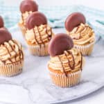 Six Peanut Butter Chocolate Chip Cupcakes with swirled frosting, drizzled with chocolate, each topped with a round chocolate-covered cookie, displayed on a marble serving tray with a blue striped cloth in the background.