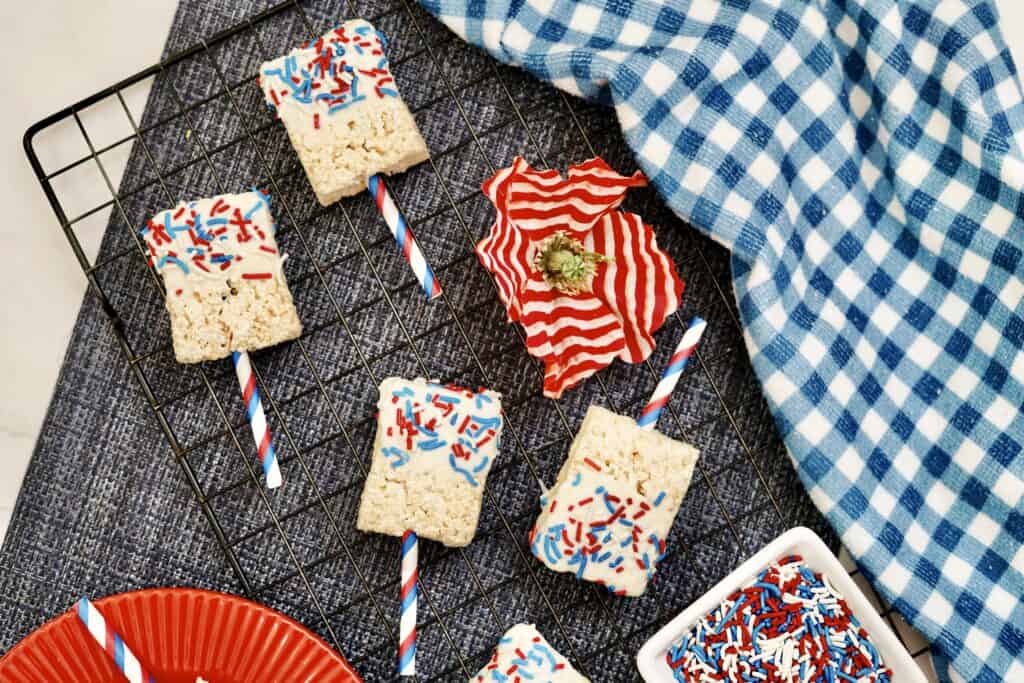 4th of July Rice Krispie treats on festive red, white, and blue striped sticks are topped with matching sprinkles and arranged on a cooling rack. A blue checkered cloth and a small bowl of sprinkles complete the scene.