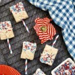 4th of July Rice Krispie treats on festive red, white, and blue striped sticks are topped with matching sprinkles and arranged on a cooling rack. A blue checkered cloth and a small bowl of sprinkles complete the scene.