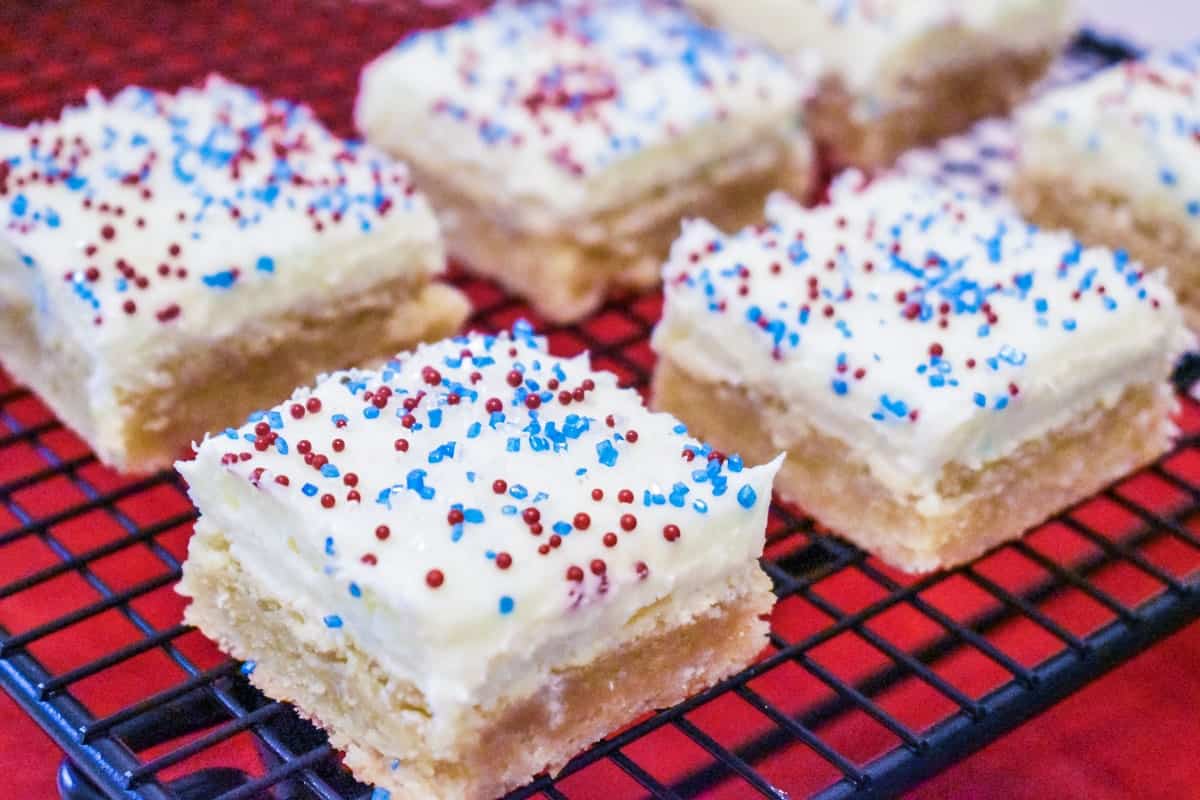Frosted sugar cookie bars topped with red, white, and blue sprinkles are cooling on a black wire rack.