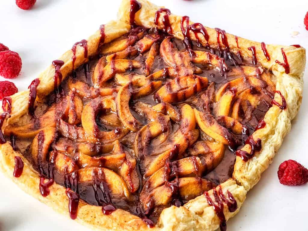 A rectangular puff pastry tart inspired by a peach tart recipe, topped with cinnamon-coated apple slices and drizzled with raspberry sauce, surrounded by fresh raspberries on a white surface.