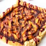 A rectangular puff pastry tart inspired by a peach tart recipe, topped with cinnamon-coated apple slices and drizzled with raspberry sauce, surrounded by fresh raspberries on a white surface.