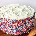 A round cake with white frosting, decorated on the sides with red, white, and blue sprinkles, sits on a wooden stand—perfect inspiration for Red White & Blue Cake Decorating Ideas.